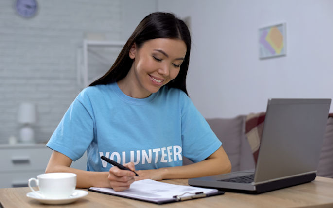 A woman sits in front of a laptop writing in a notebook with a pale blue t-shirt on that says volunteer