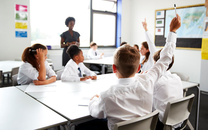 A classroom of children with their hands up and a teacher stood at the back