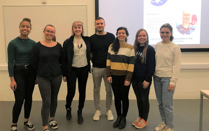 Students and invited speakers stand in a line in front of a projector after a guest lecture