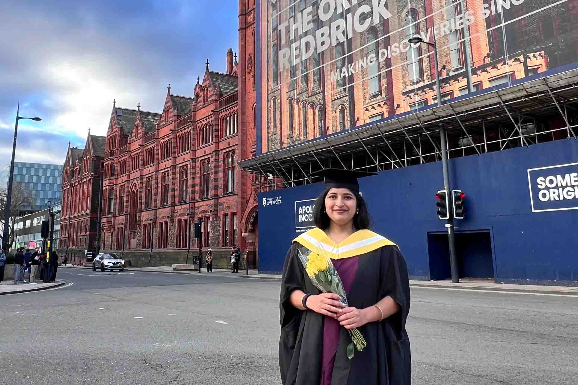graduate in academic dress in front of a red brick building