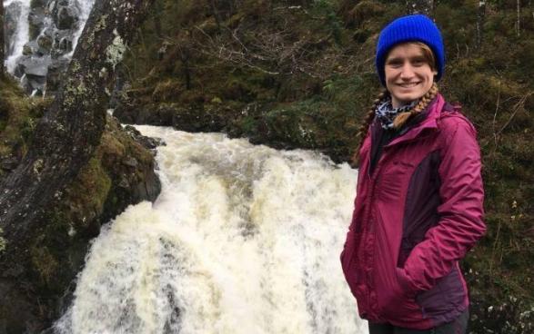 Elyse standing to the right of the frame smiling, in front of a waterfall