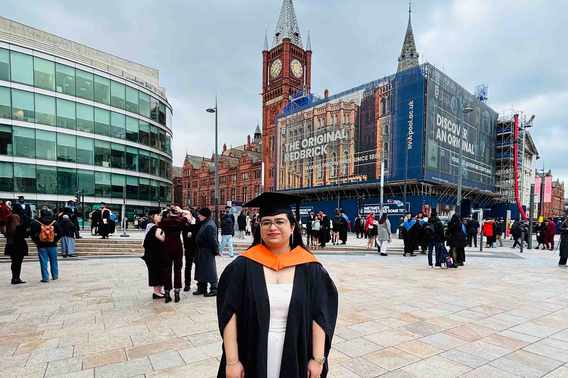 graduate in academic dress in front of a red brick building