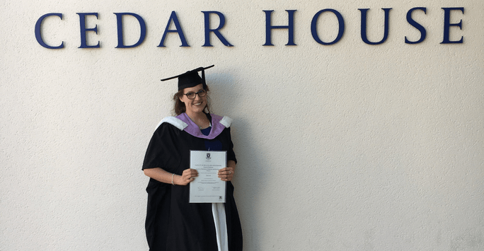 Kaylea posing with her degree certificate in front of the Cedar House sign