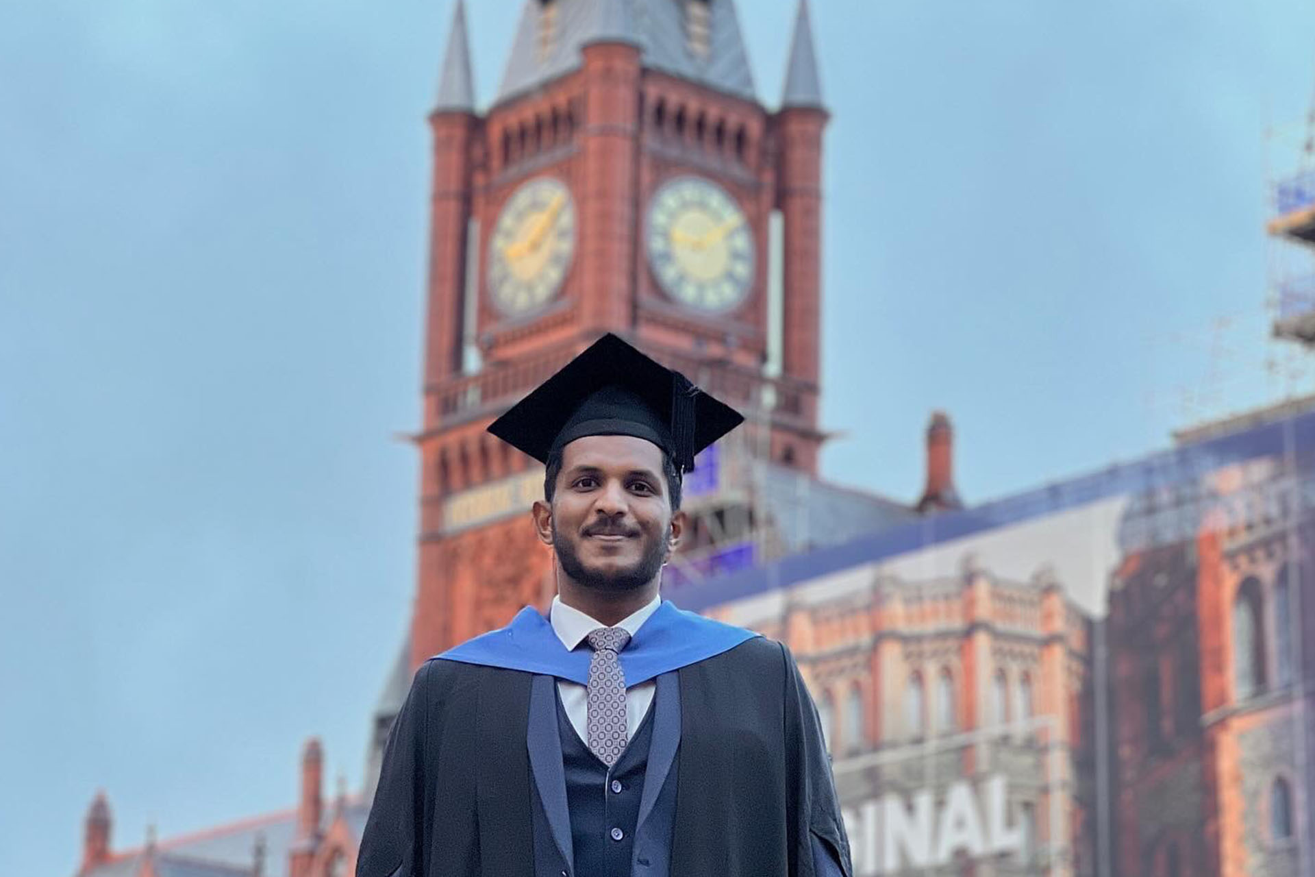 graduate in academic dress in front of a red brick building