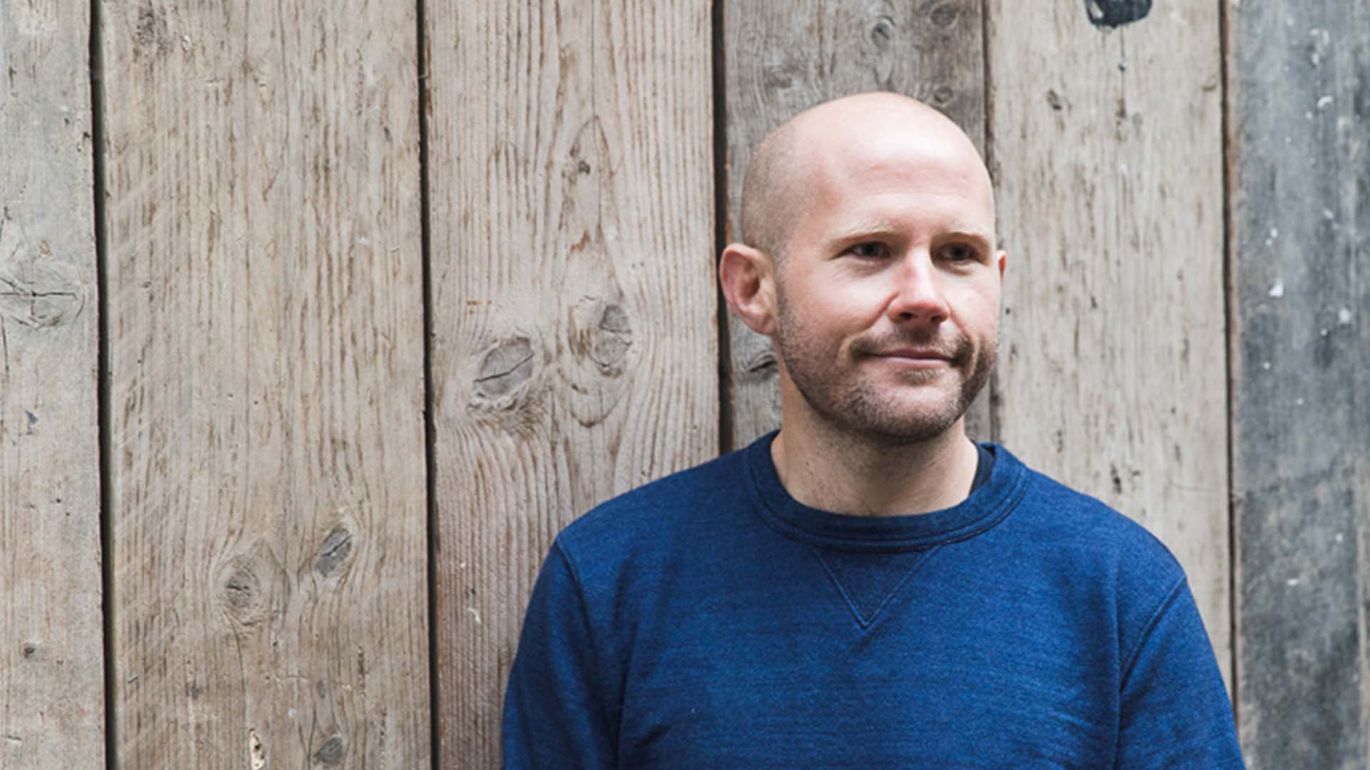 Image of person with a blue jumper on stood in front of a wooden wall